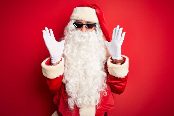Hombre de mediana edad con traje de Santa Claus y gafas de sol sobre fondo rojo aislado loco y loco gritando y gritando con expresión agresiva y los brazos levantados. Concepto de frustración
. - Foto, Imagen