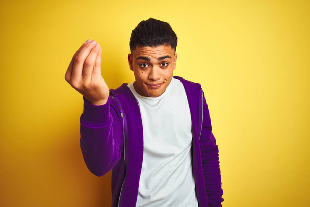 Jonge Braziliaanse man draagt paarse sweatshirt staande over geïsoleerde gele achtergrond Italiaanse gebaar met de hand en vingers zelfverzekerde uitdrukking - Foto, afbeelding