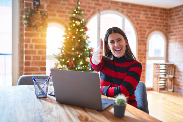 クリスマスツリーの周りの自宅でラップトップで作業テーブルに座っている美しい女性は、挨拶や歓迎として握手を提供しています。成功事業. - 写真・画像