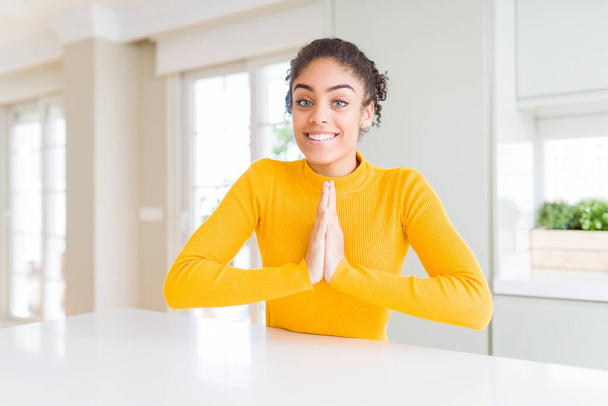Красивая африканская американка с афроволосами в обычном желтом свитере молится вместе с руками, прося прощения, уверенно улыбаясь
. - Фото, изображение