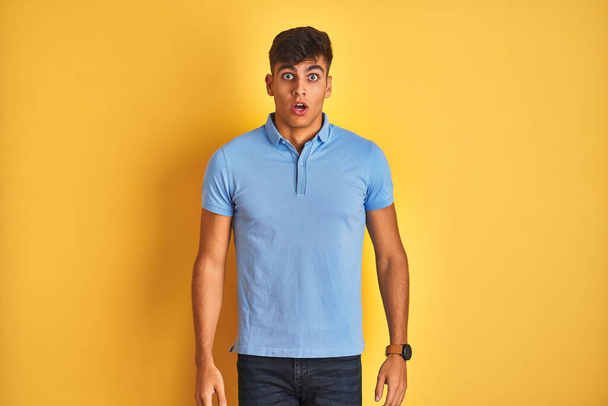 孤立した黄色の背景の上に立つ青いポロを身に着けた若いインド人男性は、驚きの表情、恐怖と興奮した顔でショックを受けました. - 写真・画像