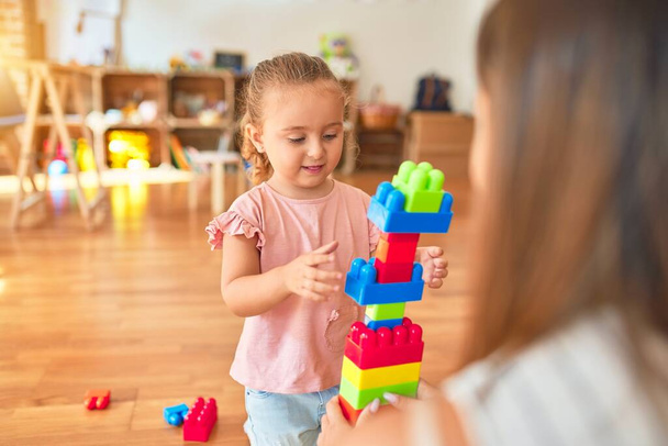 Schöne Lehrerin und blondes Kleinkind bauen Turm aus Plastikklötzen im Kindergarten - Foto, Bild