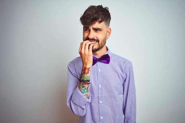 Молодий чоловік з татуюванням у фіолетовій сорочці та краватці на ізольованому білому тлі виглядає напруженим та нервовим з руками на роті кусає цвяхи. Проблема тривоги
. - Фото, зображення