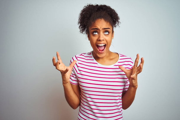 Mujer afroamericana vistiendo casual camiseta de rayas rosadas sobre aislado fondo blanco loco y loco gritando y gritando con expresión agresiva y los brazos levantados. Concepto de frustración
. - Foto, imagen