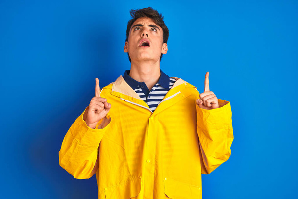 Έφηβος ψαράς αγόρι φορώντας κίτρινο αδιάβροχο πάνω από απομονωμένο φόντο έκπληκτος και έκπληκτος κοιτάζοντας προς τα πάνω και δείχνοντας με τα δάχτυλα και σήκωσε τα χέρια. - Φωτογραφία, εικόνα