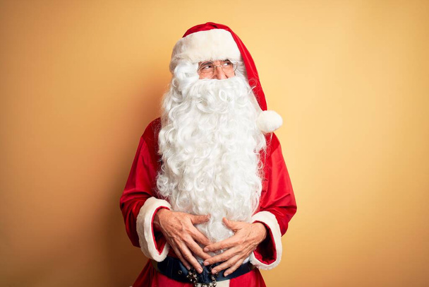 Ein gutaussehender Mann mittleren Alters im Weihnachtsmannkostüm steht vor isoliertem gelben Hintergrund und blickt mit einem Lächeln im Gesicht und natürlichem Ausdruck zur Seite. Lachen selbstbewusst. - Foto, Bild