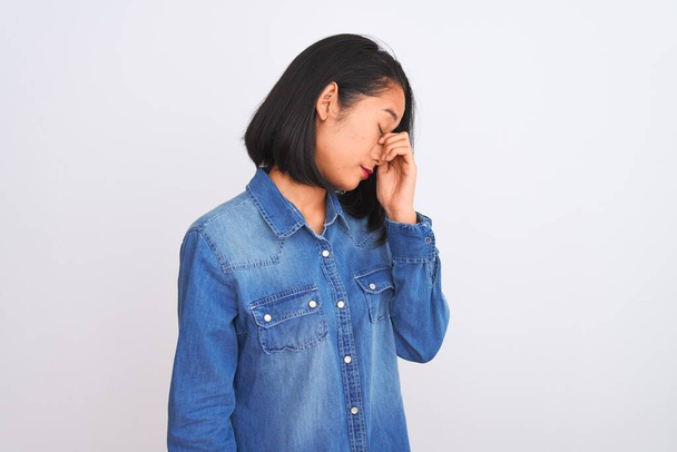Jovem mulher chinesa bonita vestindo camisa jeans em pé sobre fundo branco isolado cansado esfregando nariz e olhos sentindo fadiga e dor de cabeça. Conceito de estresse e frustração
. - Foto, Imagem