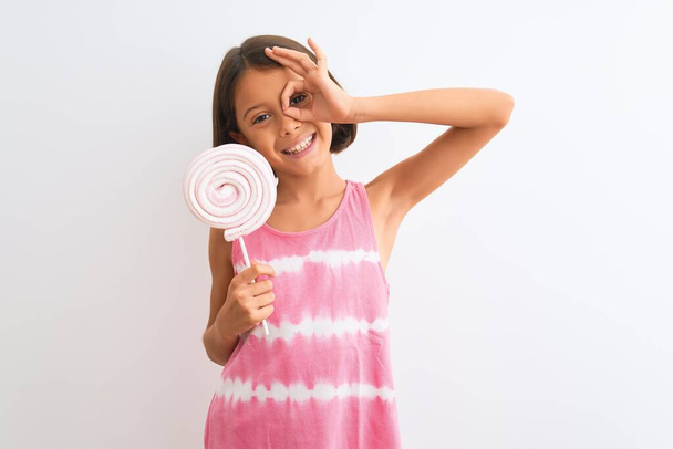Piękne dziecko dziewczyna jedzenie słodki lizak stojący nad odizolowanym białym tle z uśmiechem szczęśliwej twarzy robi ok znak z ręki na oko patrząc przez palce - Zdjęcie, obraz