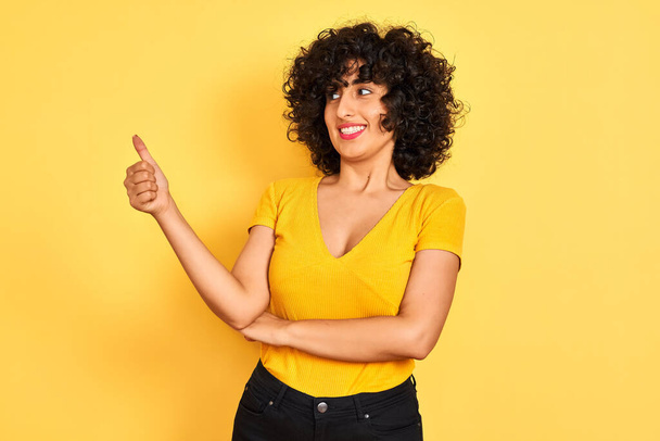 Young arab woman with curly hair wearing t-shirt standing over isolated yellow background Looking proud, smiling doing thumbs up gesture to the side - Photo, Image