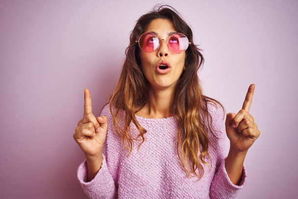 Young beautiful woman wearing fashion sunglasses standing over pink isolated background amazed and surprised looking up and pointing with fingers and raised arms. - Photo, Image