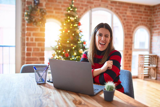 美しい女性座っているテーブルで作業ノートパソコンとともにホーム周りクリスマスツリーウインク見てカメラでセクシー式,陽気で幸せな顔. - 写真・画像