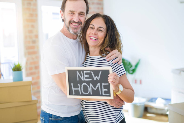 Seniorenpaar mittleren Alters, das in ein neues Haus zieht, glücklich verliebt lächelnd mit einer Tafel mit neuem Heimtext in der Hand - Foto, Bild