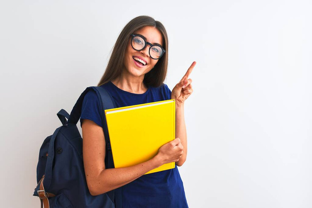 jonge student vrouw dragen rugzak bril houden boek over geïsoleerde witte achtergrond zeer gelukkig wijzend met hand en vinger naar de zijkant - Foto, afbeelding