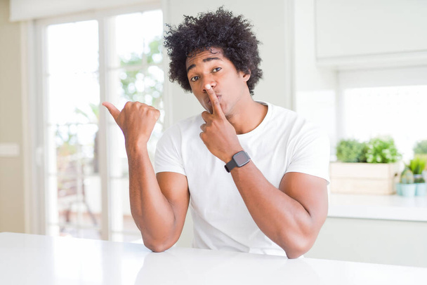 Giovane uomo afroamericano che indossa casual t-shirt bianca seduto a casa chiedendo di essere tranquillo con il dito sulle labbra che puntano con la mano verso il lato. Silenzio e concetto segreto
. - Foto, immagini
