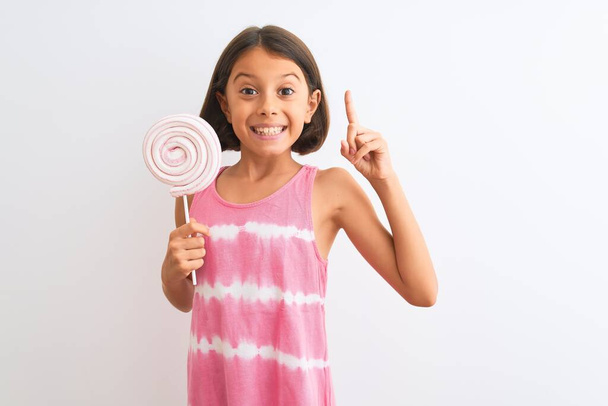 Красива дівчинка їсть солодкий льодяник, що стоїть над ізольованим білим тлом, здивована ідеєю або питанням, що вказує палець з щасливим обличчям, номер один
 - Фото, зображення