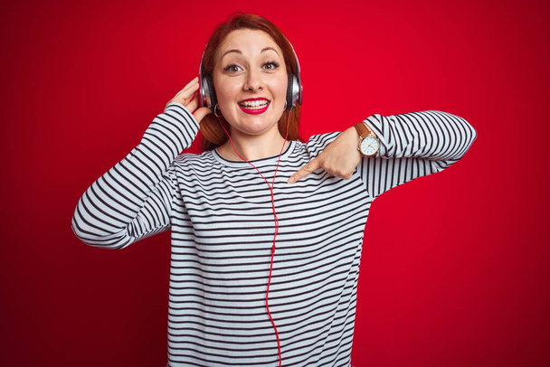 jonge roodharige vrouw luisteren naar muziek met behulp van koptelefoon over rood geïsoleerde achtergrond met verrassing gezicht wijzend vinger naar zichzelf - Foto, afbeelding
