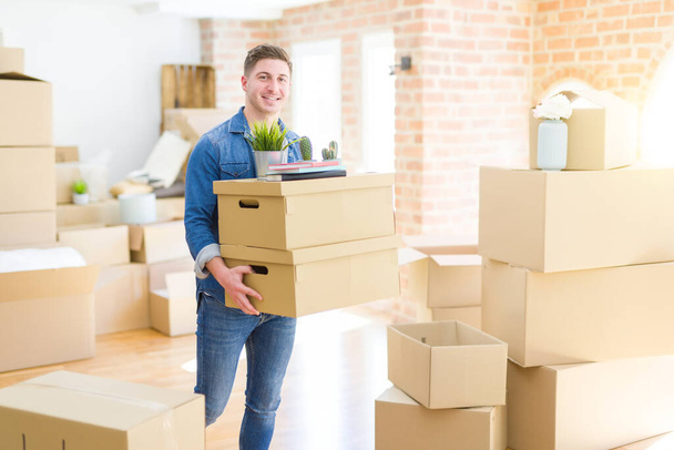 Joven guapo sonriendo feliz mudándose a un nuevo hogar, muy emocionado sosteniendo cajas de cartón en nuevo apartamento
 - Foto, Imagen