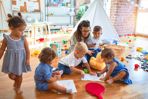 Όμορφη δασκάλα και ομάδα μικρών παιδιών που κάθονται στο πάτωμα ζωγραφίζοντας χρησιμοποιώντας χαρτί και μολύβι γύρω από πολλά παιχνίδια στο νηπιαγωγείο - Φωτογραφία, εικόνα