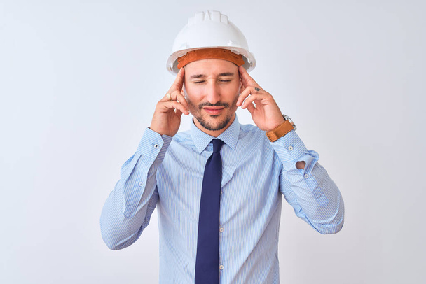 Молодой бизнесмен в защитном шлеме подрядчика на изолированном фоне с рукой на голове от боли в голове из-за стресса. Страдания от мигрени
. - Фото, изображение