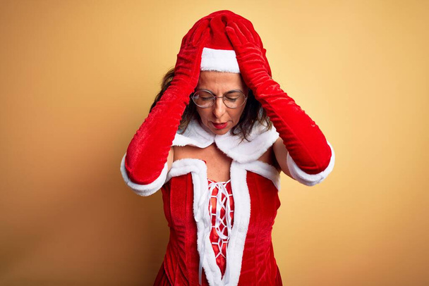 Piękna kobieta w średnim wieku ubrana w kostium Świętego Mikołaja na odizolowanym żółtym tle cierpiąca na ból głowy, zdesperowana i zestresowana, ponieważ ból i migrena. Ręce na głowę.. - Zdjęcie, obraz