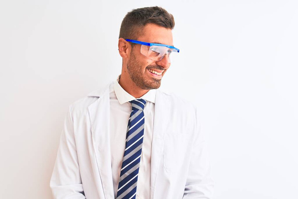Młody przystojny naukowiec noszący okulary ochronne na odosobnionym tle, patrzący z uśmiechem na twarzy, naturalnym wyrazem twarzy. Śmiejąc się pewnie. - Zdjęcie, obraz