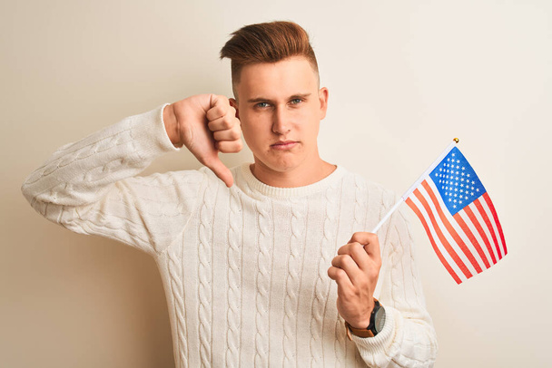 Jeune homme beau tenant le drapeau des États-Unis sur fond blanc isolé avec le visage en colère, signe négatif montrant aversion avec les pouces vers le bas, concept de rejet
 - Photo, image