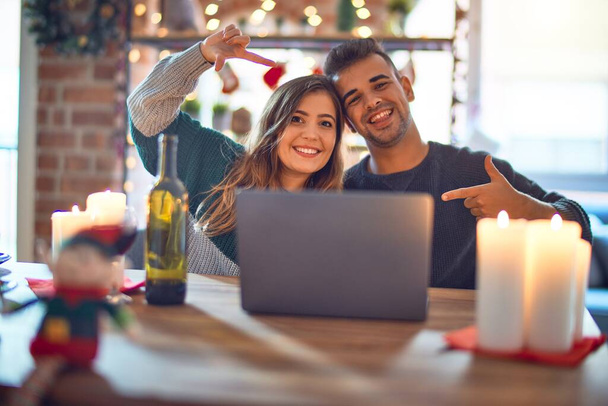 Молода красива пара сидить за допомогою ноутбука навколо різдвяної прикраси вдома, посміхаючись роблячи рамку руками і пальцями з щасливим обличчям. Концепція творчості та фотографії
. - Фото, зображення
