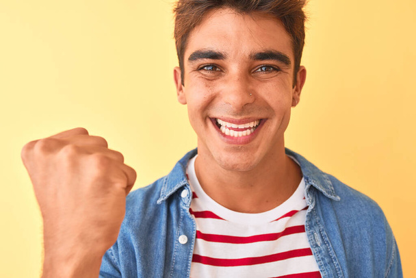 Homem bonito jovem vestindo camiseta listrada e camisa jeans sobre fundo amarelo isolado gritando orgulhoso e celebrando vitória e sucesso muito animado, torcendo emoção
 - Foto, Imagem