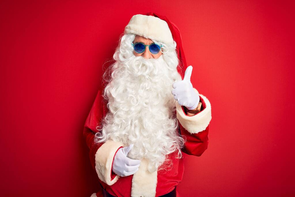 Orta yaşlı adam, Noel Baba kostümü ve güneş gözlüğü takıp izole edilmiş kırmızı arka planda mutlu bir el hareketi yapıyor. Kameraya bakıp başarıyı gösteren ifade onaylanıyor. - Fotoğraf, Görsel