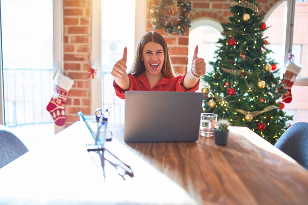 クリスマスツリーの周りの自宅でラップトップで作業テーブルに座っている美しい女性は、手で積極的なジェスチャーを行うことを承認し、親指を笑顔と成功のために幸せ。勝者のジェスチャー. - 写真・画像