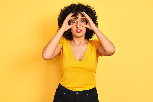 Young arab woman with curly hair wearing t-shirt standing over isolated yellow background Trying to open eyes with fingers, sleepy and tired for morning fatigue - Photo, Image
