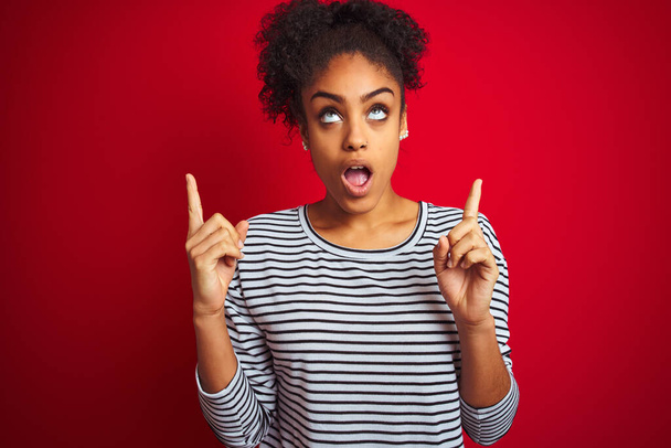 donna afroamericana che indossa navy a righe t-shirt in piedi sopra isolato sfondo rosso stupito e sorpreso guardando in alto e puntando con le dita e le braccia alzate
. - Foto, immagini