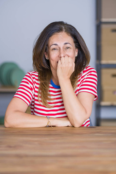 Keski-ikäinen vanhempi nainen istuu pöydässä kotona näköinen stressaantunut ja hermostunut kädet suulla puree kynnet. Ahdistuneisuusongelma
. - Valokuva, kuva