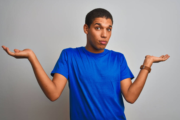 Jeune homme arabe beau portant t-shirt bleu debout sur fond blanc isolé expression inutile et confus avec les bras et les mains levées. Concept de doute
. - Photo, image