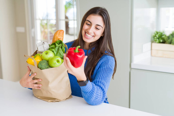 Νεαρή όμορφη γυναίκα χαμογελά κρατώντας μια χάρτινη σακούλα γεμάτη λαχανικά στο σπίτι - Φωτογραφία, εικόνα
