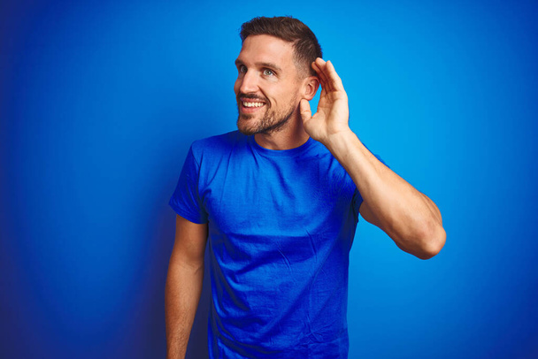 Young handsome man wearing casual t-shirt over blue isolated background smiling with hand over ear listening an hearing to rumor or gossip. Deafness concept. - Foto, afbeelding