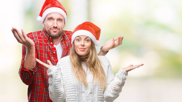 恋のクリスマス帽子をかぶっている若いカップルは、腕と手を上げて背景無知と混乱式を分離しました。疑いのコンセプト. - 写真・画像