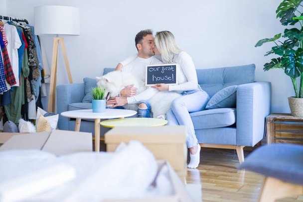 Young beautiful couple with dog sitting on the sofa holding blackboard with message kissing at new home around cardboard boxes - Foto, Imagen