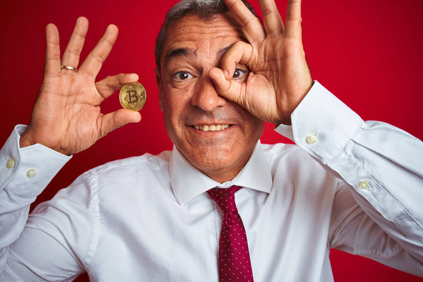 Jóképű középkorú férfi kezében bitcoin felett áll elszigetelt piros háttér boldog arc mosolygós csinál rendben jel kéz a szemen néz keresztül ujjait - Fotó, kép