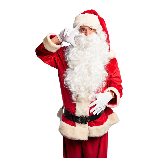 Красивый мужчина средних лет в костюме Санта-Клауса и с бородой стоя делает символ мира с пальцами на лице, улыбаясь весело показывая победу
 - Фото, изображение