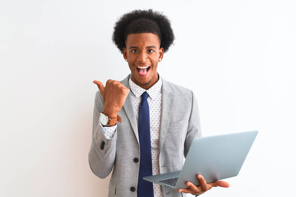 Fiatal afro-amerikai üzletember laptoppal áll elszigetelt fehér háttér mutatva, és mutatja hüvelykujjával oldalra boldog arc mosolygós - Fotó, kép