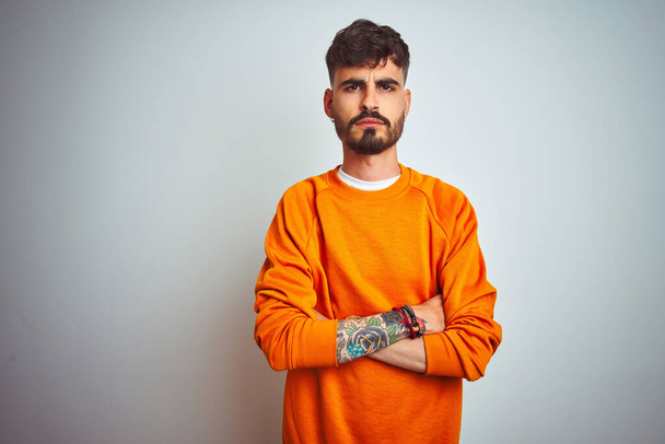 Joven hombre con tatuaje usando suéter naranja de pie sobre fondo blanco aislado escéptico y nervioso, desaprobando la expresión en la cara con los brazos cruzados. Persona negativa
. - Foto, imagen