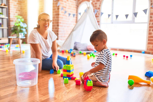 Beautiful teacher and toddler boy playing with construction blocks bulding tower at kindergarten - Photo, image