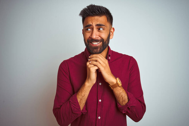 Jeune homme indien portant chemise élégante rouge debout sur fond gris isolé riant nerveux et excité avec les mains sur le menton regardant vers le côté
 - Photo, image