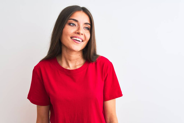 Jonge mooie vrouw draagt rode casual t-shirt staan over geïsoleerde witte achtergrond weg te kijken naar kant met glimlach op het gezicht, natuurlijke expressie. Lachend vol vertrouwen. - Foto, afbeelding
