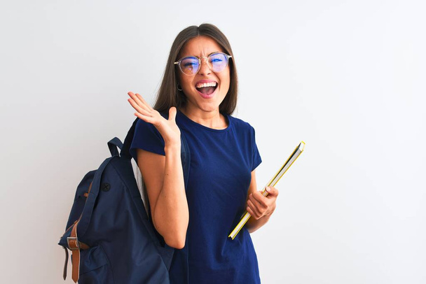Young student woman wearing backpack glasses holding book over isolated white background very happy and excited, winner expression celebrating victory screaming with big smile and raised hands - Foto, immagini