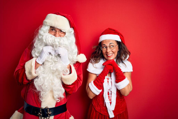 Middle age couple wearing Santa costume and glasses over isolated red background laughing nervous and excited with hands on chin looking to the side - Photo, Image