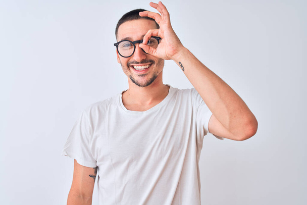Young handsome man wearing glasses and standing over isolated background doing ok gesture with hand smiling, eye looking through fingers with happy face. - Фото, зображення
