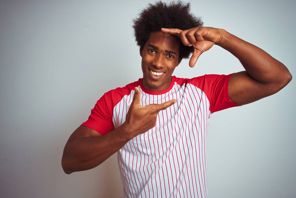 Afrikanisch-amerikanischer Mann mit Afro-Haaren, der ein rot gestreiftes T-Shirt über isoliertem weißem Hintergrund trägt und lächelnd mit Händen und Fingern ein glückliches Gesicht macht. Kreativität und Fotografiekonzept. - Foto, Bild