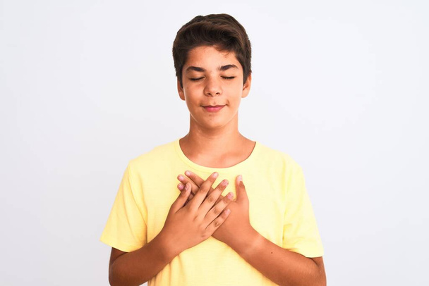 gutaussehender Teenager, der vor weißem Hintergrund steht und mit geschlossenen Augen und dankbarer Geste auf dem Gesicht lächelnd die Hände auf der Brust hält. Gesundheitskonzept. - Foto, Bild